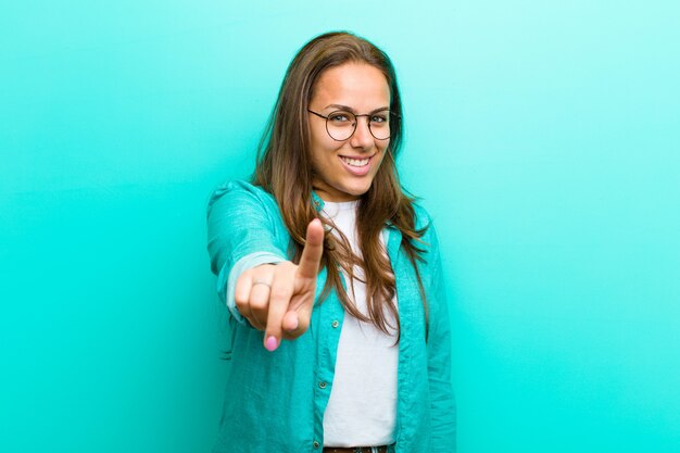 Jeune femme souriante et fière de faire la pose numéro un triomphalement, se sentant comme un leader contre le mur bleu