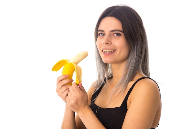 Jeune femme souriante en haut noir mangeant une banane sur fond blanc en studio