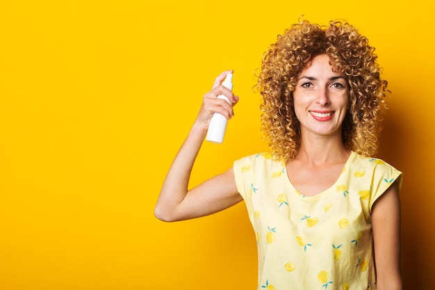 Une jeune femme souriante hydrate ses cheveux avec un spray sur fond jaune