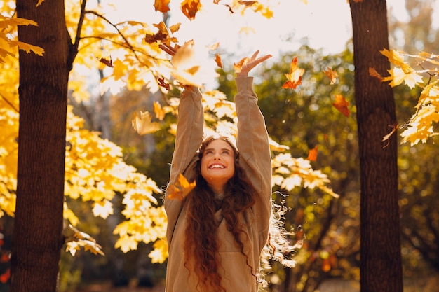 Une jeune femme souriante jette les feuilles d'automne dans la forêt au coucher du soleil