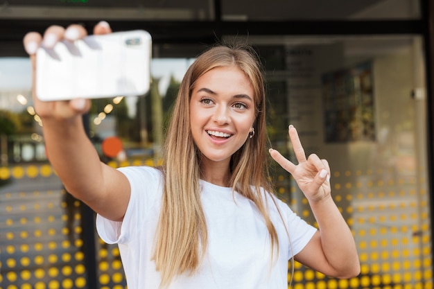 Jeune femme souriante et joyeuse à l'extérieur dans la rue près du centre d'affaires à l'aide d'un téléphone portable prend un selfie montrant la paix.