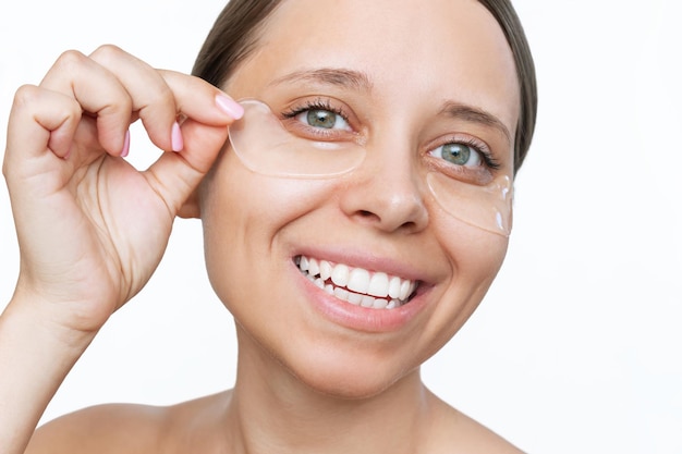 Une jeune femme souriante et joyeuse met des patchs transparents incolores en gel sur la peau sous ses yeux