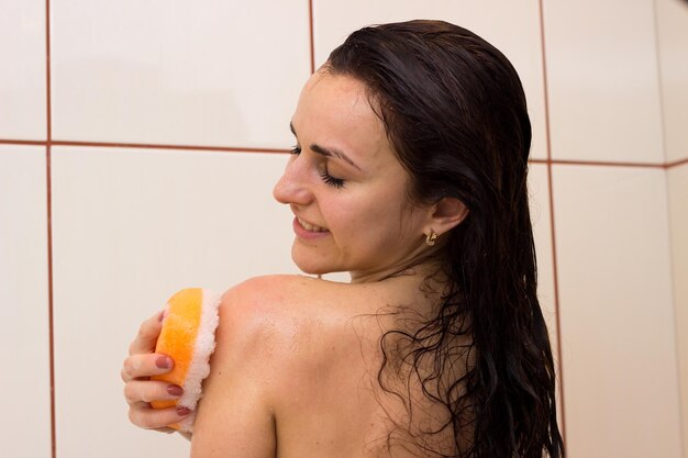 Jeune femme souriante avec de longs cheveux noirs mouillés se lavant avec une éponge orange dans sa salle de bain lumineuse