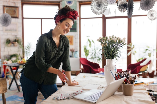 Jeune femme souriante magnifique aux cheveux bouclés noirs s'appuyant sur la table regardant joyeusement dans un ordinateur portable avec des croquis autour de passer du temps dans un atelier confortable moderne avec de grandes fenêtres