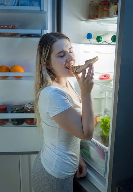 Jeune femme souriante mangeant de la pizza dans la cuisine la nuit