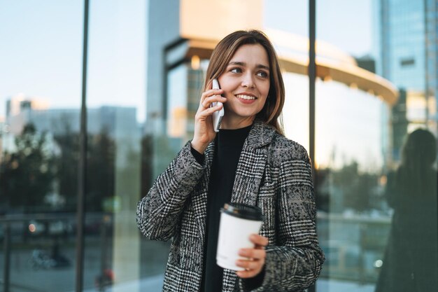 Jeune femme souriante en manteau avec une tasse de café à l'aide d'un téléphone portable dans la rue de la ville en soirée