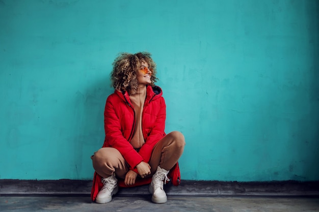 Jeune femme souriante à la mode aux cheveux bouclés, en veste rouge accroupie devant le mur et en détournant les yeux.