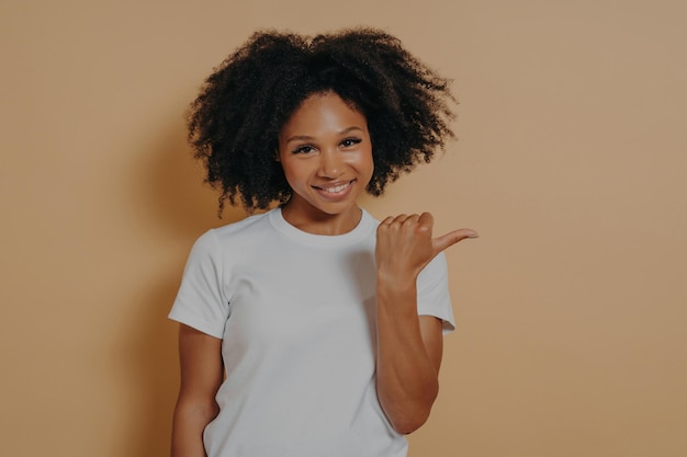 Jeune femme souriante à la peau foncée, cheveux bouclés, pointant de côté avec le pouce isolé sur un mur beige