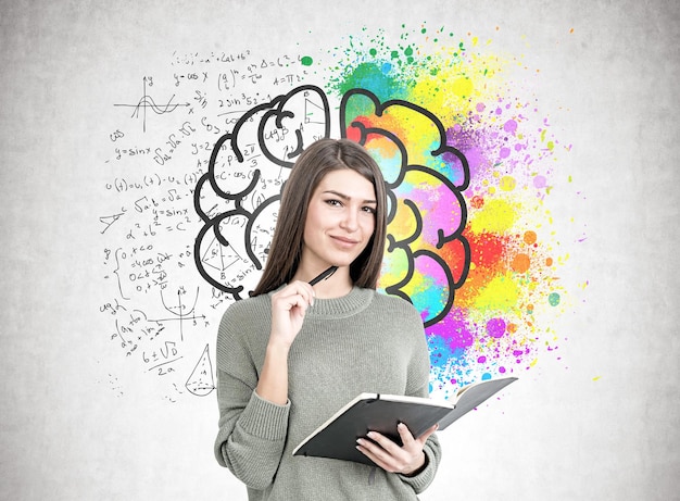 Une jeune femme souriante portant un chandail gris tient un planificateur et un stylo. Un mur de béton avec un croquis de cerveau coloré