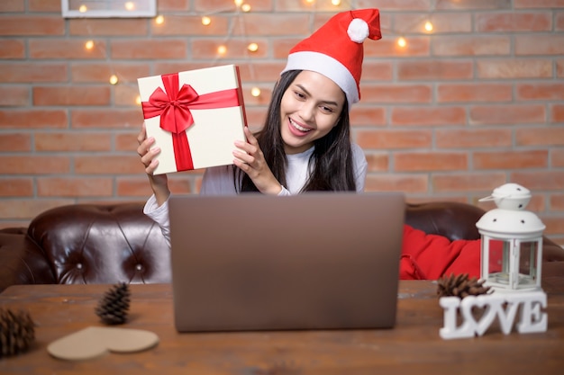 Une jeune femme souriante portant un chapeau de père Noël rouge faisant un appel vidéo sur le réseau social avec la famille et les amis le jour de Noël.