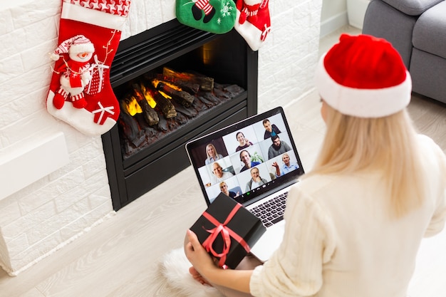 Une jeune femme souriante portant un chapeau de père Noël rouge faisant un appel vidéo sur un réseau social avec sa famille et ses amis le jour de Noël.