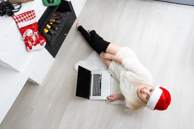 Une jeune femme souriante portant un chapeau de père Noël rouge faisant un appel vidéo sur un réseau social avec sa famille et ses amis le jour de Noël.