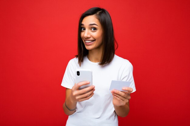 Jeune femme souriante portant des vêtements de tous les jours isolés sur fond tenant un téléphone et une carte de crédit payant des achats en ligne par carte de crédit en regardant la caméra