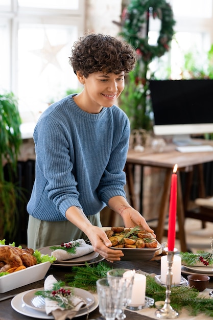 Jeune femme souriante en pull bleu et pantalon gris portant un bol avec des pommes de terre au four faites maison et le mettant sur une table de fête