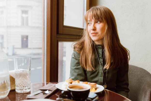 Jeune femme souriante et réfléchie regardant loin dans un café tout en buvant du café et en mangeant du gâteau au fromage