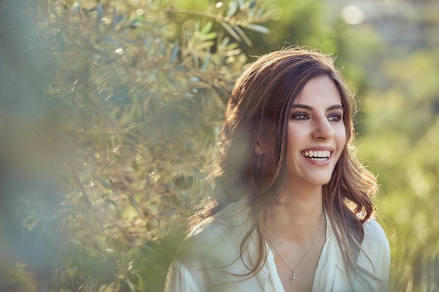 Jeune femme souriante regardant au loin avec le sourire tout en se tenant près d'un arbre vert luxuriant dans la campagne sur un arrière-plan flou le jour de l'été