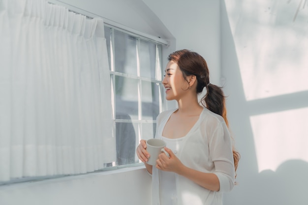 Jeune femme souriante regardant par la fenêtre avec une tasse de boisson