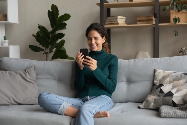 Une jeune femme souriante se détend sur le canapé avec son smartphone, navigue dans les applications de son appareil mobile, regarde l'écran de son téléphone, discute ou fait des achats en ligne, profite de son temps libre avec son gadget, regarde des vidéos drôles.