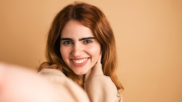 Une jeune femme souriante se fait un selfie dans un studio beige.