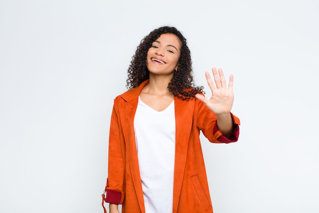 Jeune femme souriante et sympathique, montrant le numéro cinq ou cinquième avec la main vers l'avant, compte à rebours sur le mur blanc