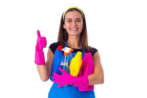 Jeune femme souriante en T-shirt bleu et tablier avec des gants roses tenant des détergents et montrant le pouce vers le haut sur fond blanc en studio