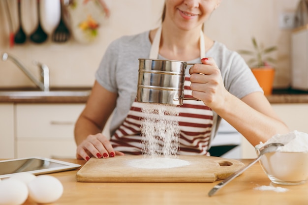 La jeune femme souriante tamise la farine avec un tamis en fer avec tablette sur la table de la cuisine. Cuisiner à la maison. Préparer la nourriture.