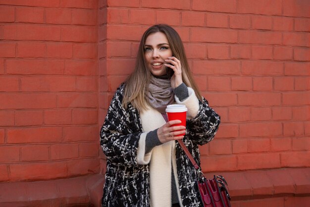 Jeune femme souriante avec téléphone et café dans une rue de la ville. Belle blonde dans un manteau élégant et chaud sur fond de mur de briques rouges. Blogs et télétravail.