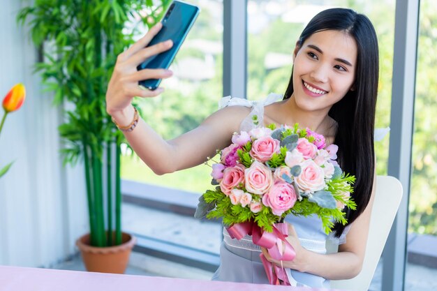 Photo une jeune femme souriante tenant des fleurs se fait un selfie avec un smartphone à la maison