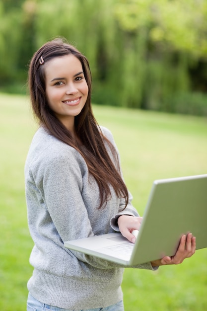 Jeune femme souriante tenant son ordinateur portable tout en se tenant debout dans un parc