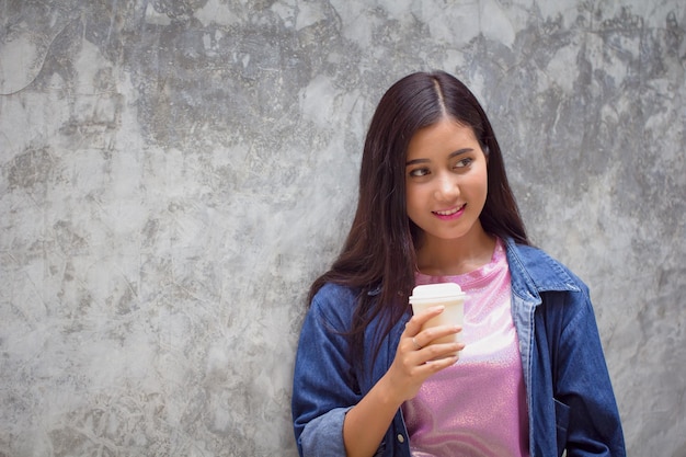 Une jeune femme souriante tenant une tasse jetable en détournant le regard contre le mur.