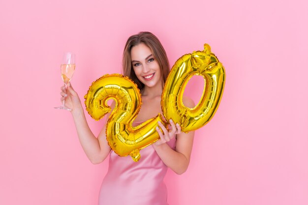 Une jeune femme souriante tient un ballon en feuille d'or et une coupe de champagne sur une fête d'anniversaire sur fond rose