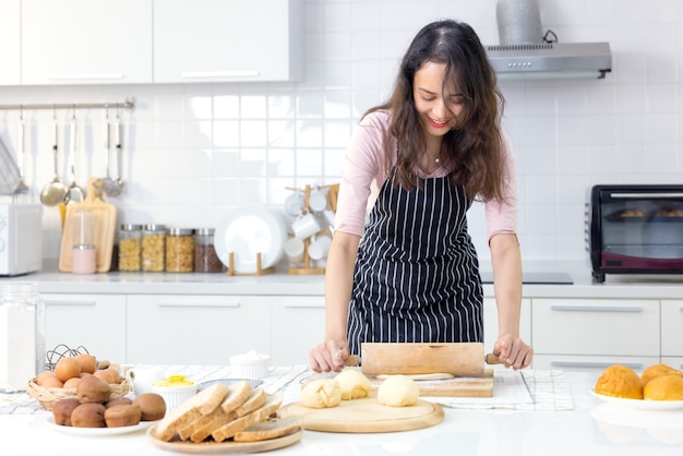 Une jeune femme souriante travaille avec une goupille en bois pour faire une tarte sucrée ou de la pâte à pâtisserie dans la cuisine, une femme aimante et heureuse du millénaire cuisinant dans un tablier préparant un dîner en famille ou des petits pains à dessert