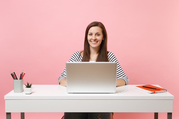 Jeune femme souriante en vêtements décontractés travaillant sur un projet avec un ordinateur portable alors qu'elle était assise au bureau