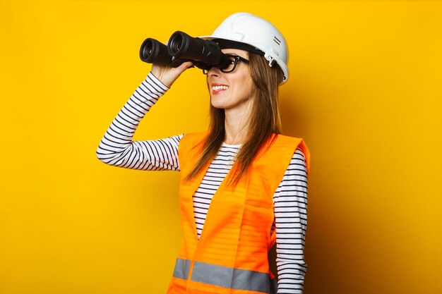 Jeune femme avec un sourire dans un gilet et un casque regarde à travers des jumelles sur un fond jaune Concept de construction nouveau bâtiment Bannière