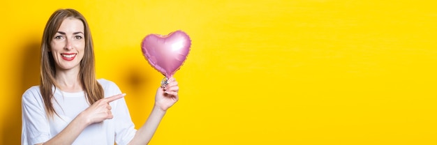 Jeune femme avec un sourire tient un ballon en forme de cœur dans ses mains et pointe son doigt dessus sur un fond jaune. Bannière.