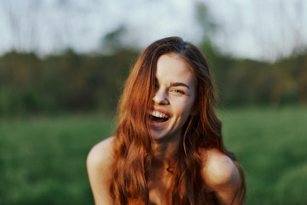 Une jeune femme sourit et regarde dans la caméra avec ses longs cheveux brillants ondulés rouges dans un parc avec de l'herbe verte au coucher du soleil d'été Le concept de beauté et de soins capillaires sains