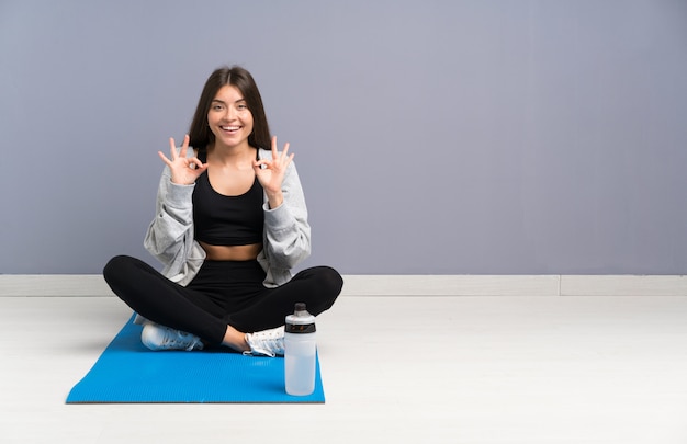 Jeune femme de sport assis sur le sol avec tapis montrant un signe ok avec les doigts