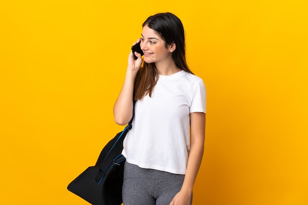 Jeune femme de sport avec sac de sport isolé sur jaune en gardant une conversation avec le téléphone mobile avec quelqu'un