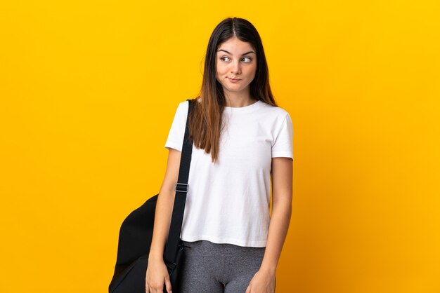 Jeune femme de sport avec sac de sport isolé sur mur jaune ayant des doutes tout en regardant