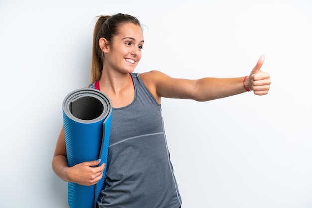 Jeune femme sportive allant à des cours de yoga tout en tenant un tapis isolé sur fond blanc en donnant un geste du pouce levé