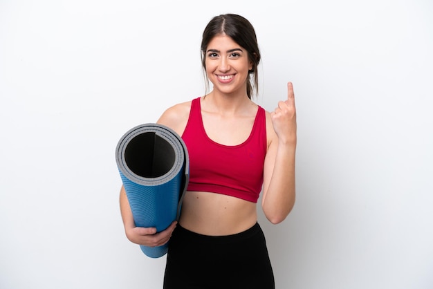 Jeune femme sportive allant à des cours de yoga tout en tenant un tapis isolé sur fond blanc montrant et levant un doigt en signe du meilleur