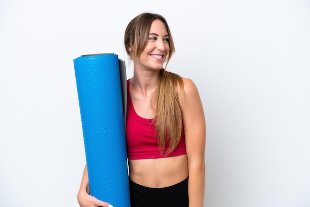 Jeune femme sportive allant à des cours de yoga tout en tenant un tapis isolé sur fond blanc regardant sur le côté et souriant