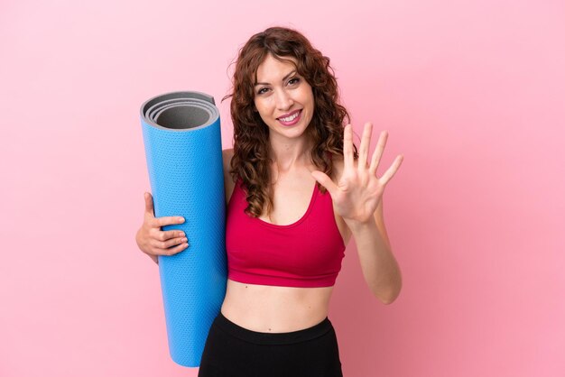 Jeune femme sportive allant à des cours de yoga tout en tenant un tapis isolé sur fond rose en comptant cinq avec les doigts