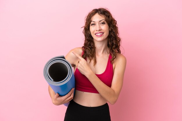 Jeune femme sportive allant à des cours de yoga tout en tenant un tapis isolé sur fond rose pointant vers le côté pour présenter un produit