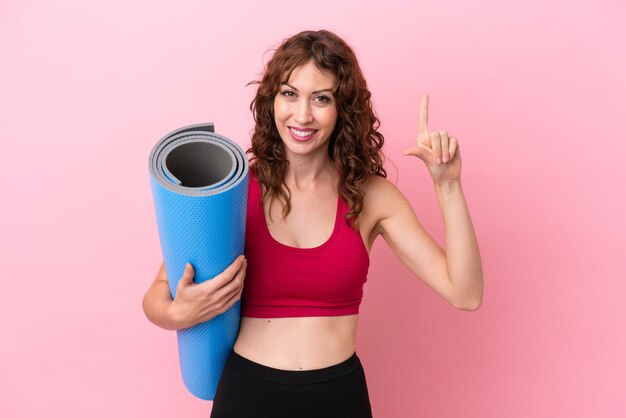 Jeune femme sportive allant à des cours de yoga tout en tenant un tapis isolé sur fond rose pointant vers une excellente idée