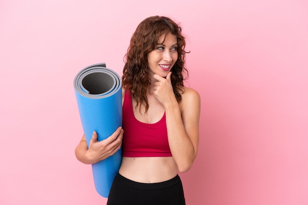 Jeune femme sportive allant à des cours de yoga tout en tenant un tapis isolé sur fond rose regardant sur le côté et souriant