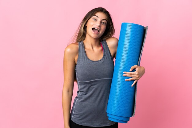 Jeune femme sportive allant à des cours de yoga tout en tenant un tapis isolé sur rose avec surprise et expression faciale choquée