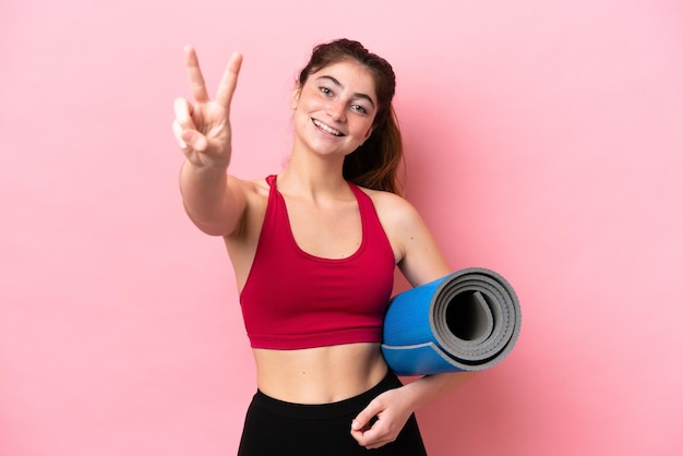 Jeune femme sportive allant à des cours de yoga tout en tenant un tapis souriant et montrant le signe de la victoire