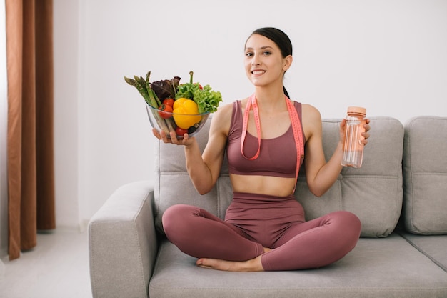 Jeune femme sportive assise sur un canapé tenant une variété de légumes une bouteille d'eau et une règle enveloppée concept de perte de poids et d'alimentation saine