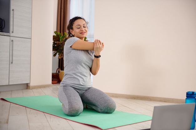 Jeune femme sportive assise sur un tapis de fitness et étirant son corps à gauche tout en travaillant à la maison pendant le verrouillage